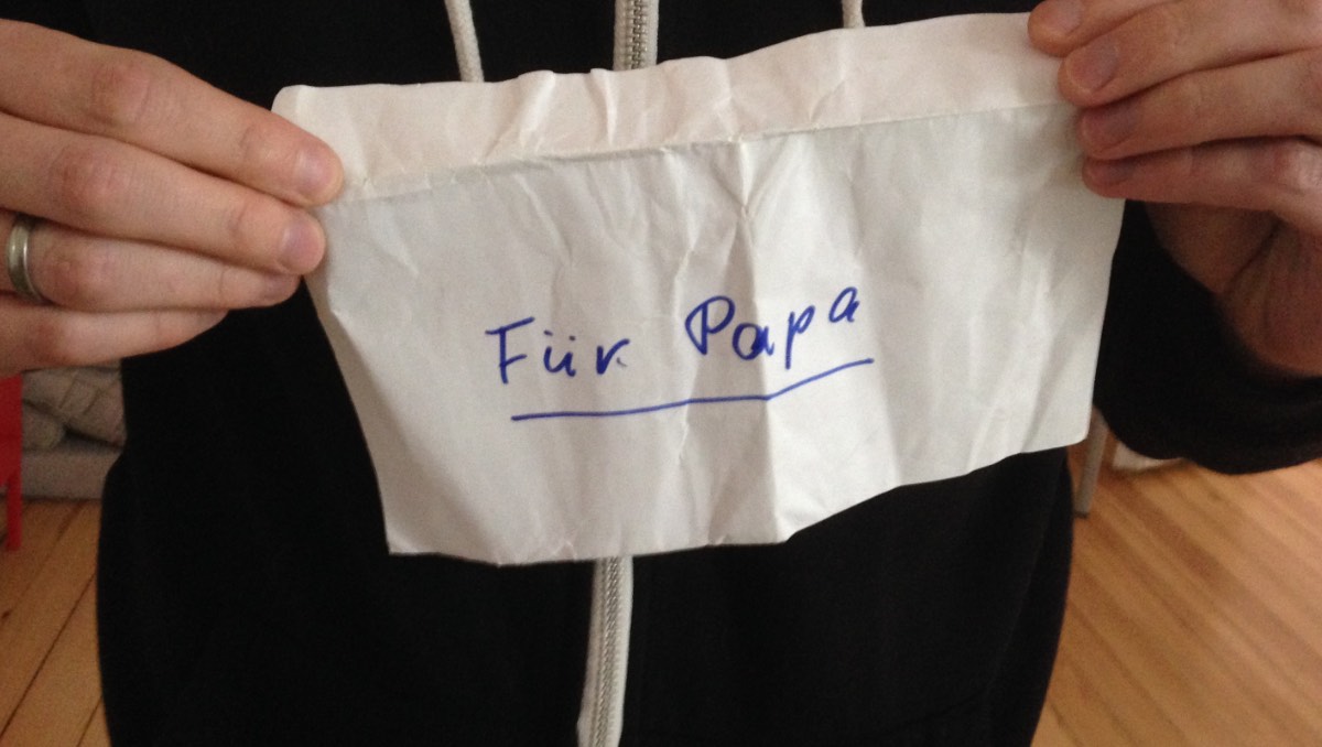 Ein Mann mit Ehering am Finger hält einen zerknitterten Briefumschlag hoch, auf dem 'für Papa' steht.