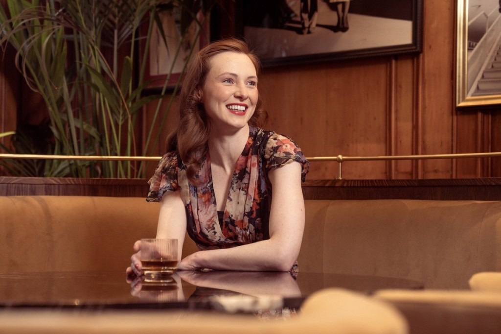 Eine junge Frau sitzt in einer Bar und hält ein gefülltes Glas in der Hand.