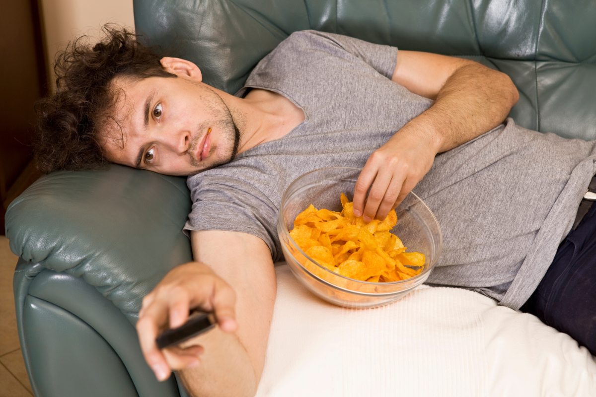 Ein fauler Mann liegt auf der Couch, schaut Fernsehen und isst Chips.