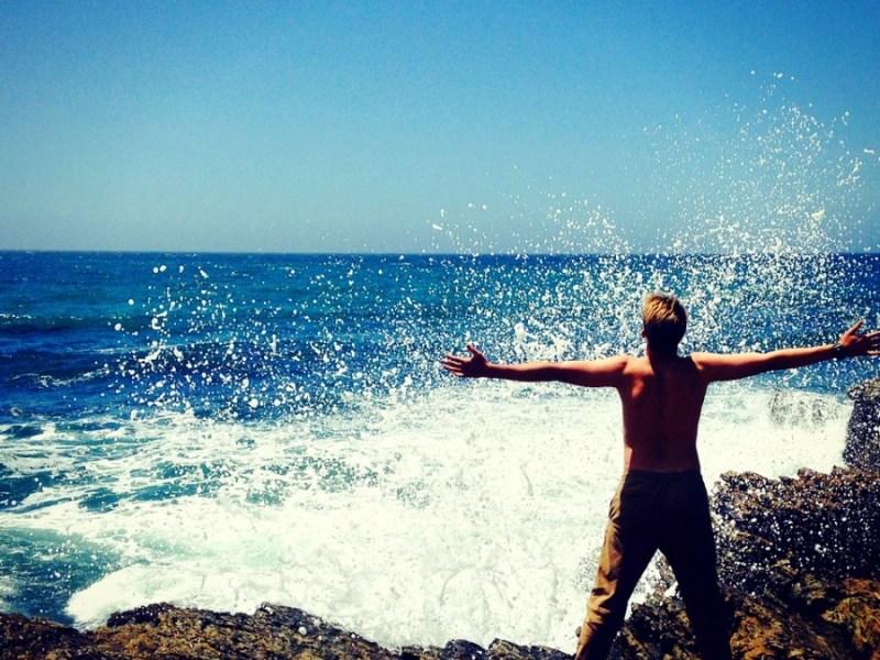 Ein Mann steht mit freiem Oberkörper und ausgebreiteten Armen am Meer.