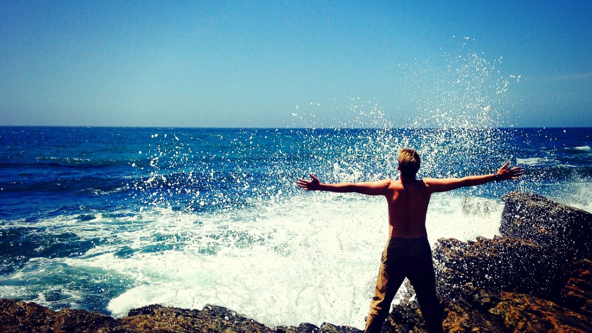 Ein Mann steht mit freiem Oberkörper und ausgebreiteten Armen am Meer.