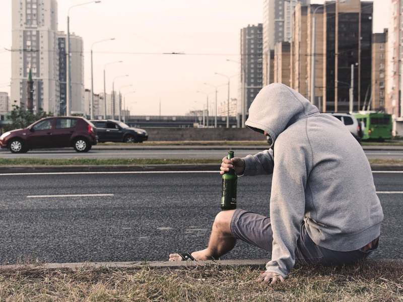 Mann mit Kapuze über Kopf sitzt mit Bierflasche am Straßenrand