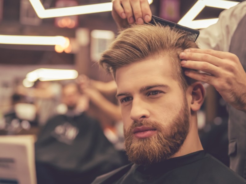 Ein gut aussehender bärtiger Mann liest eine Zeitung, während er sich vom Friseur die Haare schneiden lässt.