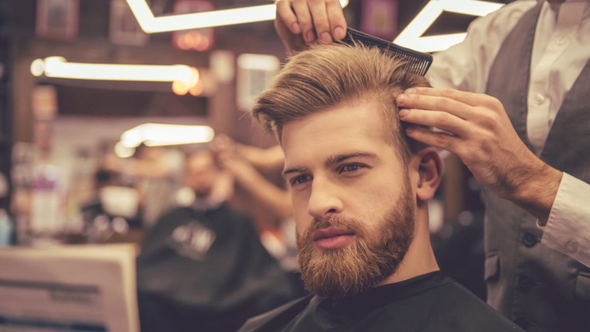 Ein gut aussehender bärtiger Mann liest eine Zeitung, während er sich vom Friseur die Haare schneiden lässt.