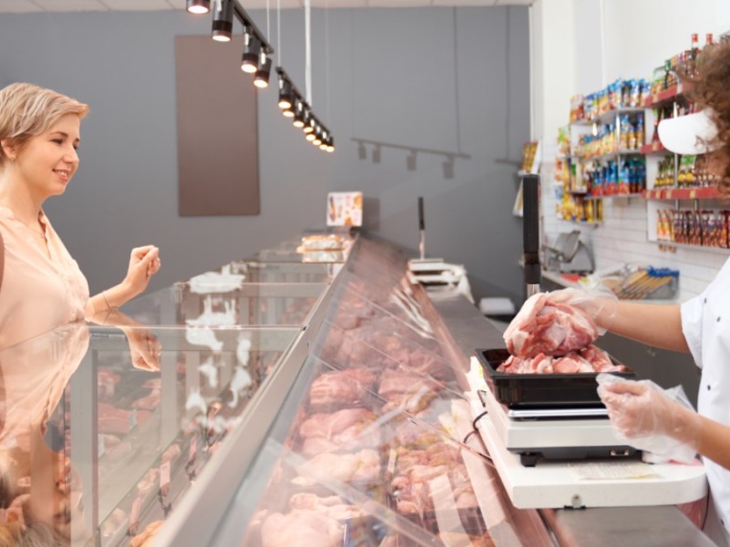 Seitenansicht einer jungen lächelnden Frau mit lockigem Haar, die einer positiven Dame rohes Schweinefleisch vorführt.